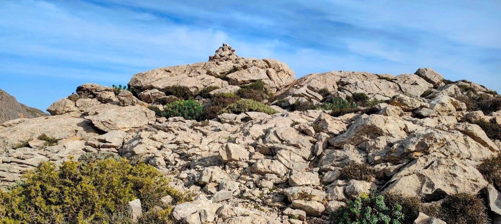 Puig del Tossals Verd desde Cúber - Caminando por Mallorca Vista de terreno rocoso con vegetación escasa y un hito en la cima de una roca, bajo un cielo despejado.