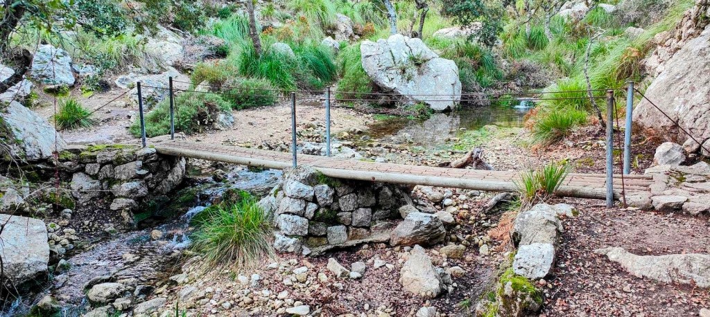 Puente de madera que cruza un arroyo en un entorno natural, rodeado de rocas y vegetación verde.