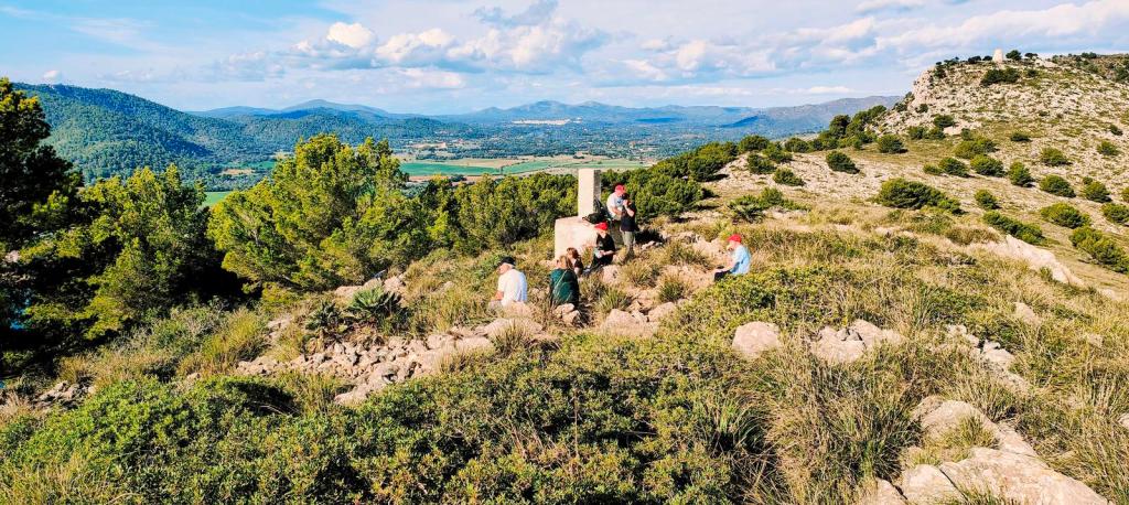 Grupo de personas sentadas alrededor de un vértice geodésico en un paisaje montañoso, con vista a un valle y colinas en el fondo.