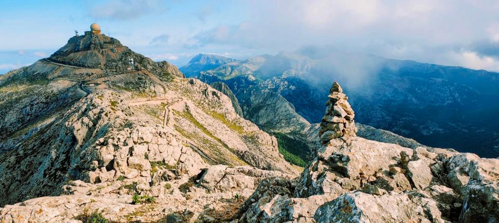 Vista panorámica desde la cima del Penyal des Migdia, mostrando un paisaje montañoso con un hito de piedras en primer plano y un radar en la cima de una montaña lejana.