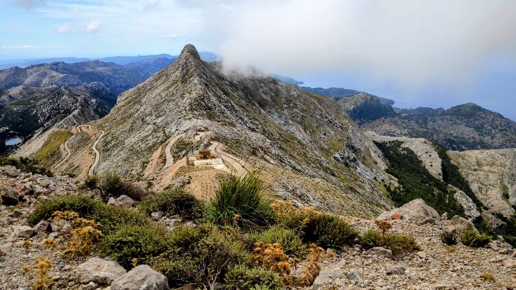 Vista panorámica del Penyal des Migdia en la Sierra de Tramuntana, Mallorca, con nubes cubriendo la cima y un sendero que serpentea por el terreno rocoso.