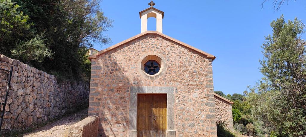 Vista de una pequeña iglesia de piedra con un campanario, ubicada en un entorno natural. El cielo es claro y se pueden ver árboles y vegetación alrededor.
