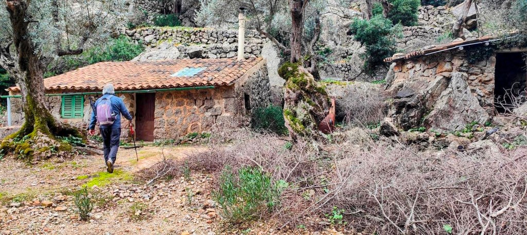 Hombre caminando hacia una casa de piedra con techo de tejas, rodeada de vegetación y árboles en un paisaje natural, en la ruta 'Puig des Moro desde Sóller'.
