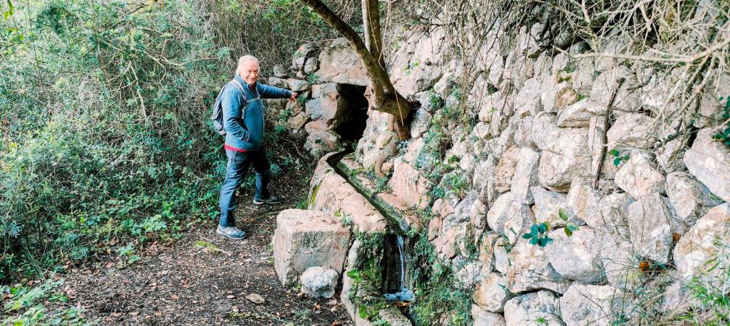 Hombre apoyado en una pared de piedra junto a una fuente, rodeado de vegetación en un sendero natural.