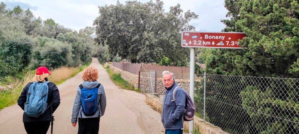 Tres personas caminando por un camino rural, con un cartel que indica la distancia y la inclinación hacia Bonany.