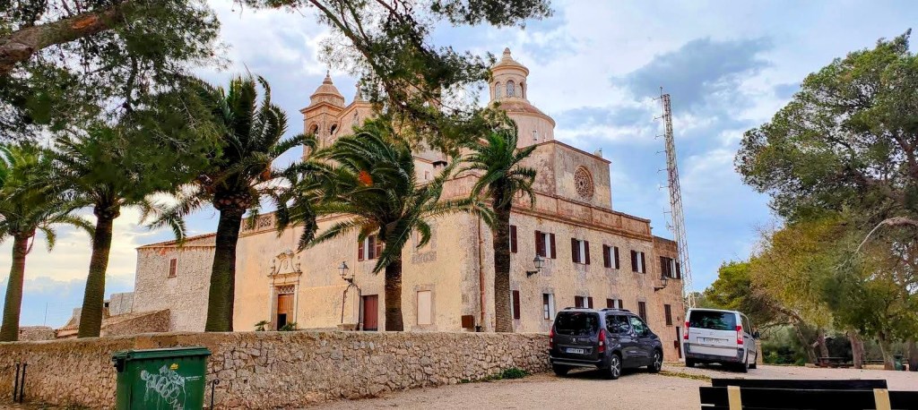 Vista del Santuario de Bonany rodeado de palmeras, con coches aparcados en el lado inferior y un cielo nublado al fondo.