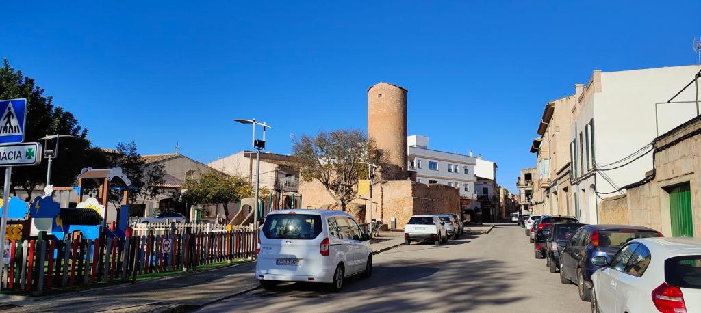 Ruta Hidráulica 3 de Porreres - Caminando por Mallorca Vista de una calle en Porreres con un parque infantil a la izquierda y un molino de viento antiguo en el fondo, bajo un cielo azul claro.