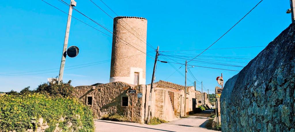 Ruta Hidráulica 3 de Porreres - Caminando por Mallorca Vista de un molino de viento tradicional de piedra con base cuadrada y torre cilíndrica, rodeado de edificios y vegetación en un paisaje rural bajo un cielo despejado.
