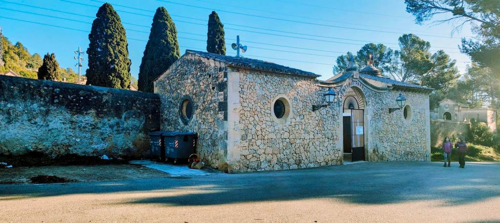 Aparcamiento del cementerio de la Vileta, con un edificio de piedra y dos personas de pie frente a él, rodeado de árboles.