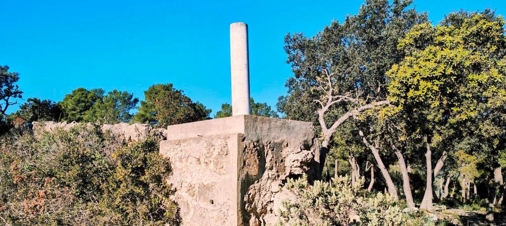 Estructura de piedra con un tubo en la parte superior, rodeada de vegetación y árboles, bajo un cielo azul claro.