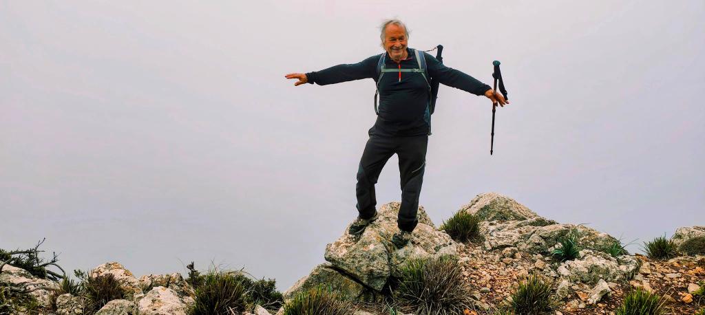 Hombre de pie sobre una roca en una cima montañosa, con los brazos extendidos, mientras sostiene un bastón de senderismo. Fondo de niebla que cubre la vista.