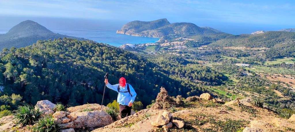 Persona con gorra roja caminando sobre rocas en una montaña, con vistas panorámicas a la costa y un valle cubierto de árboles.