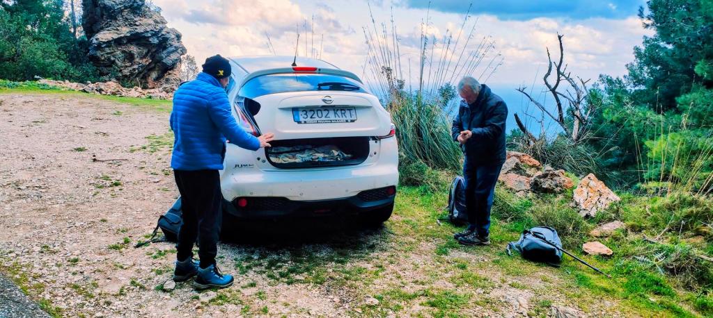 Dos personas en un área natural, una frente al maletero de un coche blanco y la otra ajustándose la ropa. El fondo muestra vegetación y rocas.