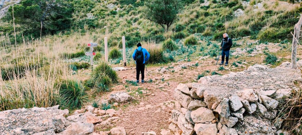 Dos senderistas observan un cruce de caminos en un paisaje montañoso con vegetación, marcadores de sendero y rocas.