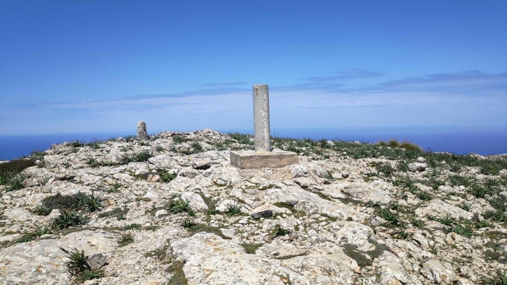 Vista desde la cima de la Mola de s'Esclop, con un vértice geodésico en primer plano, rodeado de rocas y vegetación en un paisaje montañoso bajo un cielo azul.