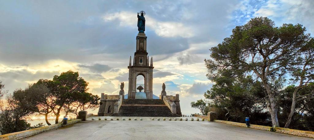 Vista del monumento a Cristo Rey en el Puig de Sant Salvador, con una estatua de bronce de Cristo y un paisaje de árboles y nubes al fondo.