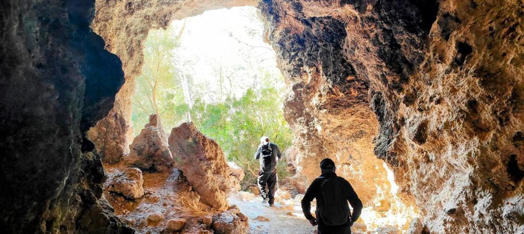 Dos personas caminan por el interior de una cueva, iluminadas por la luz que entra en el fondo. Las paredes de la cueva son rocosas y hay vegetación visible al exterior.