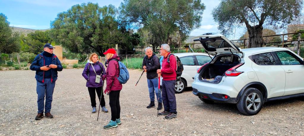 Grupo de caminantes preparándose para iniciar la ruta "Puig de la Tudossa y Puig del Corb" en un aparcamiento rural, rodeados de árboles y vehículos.