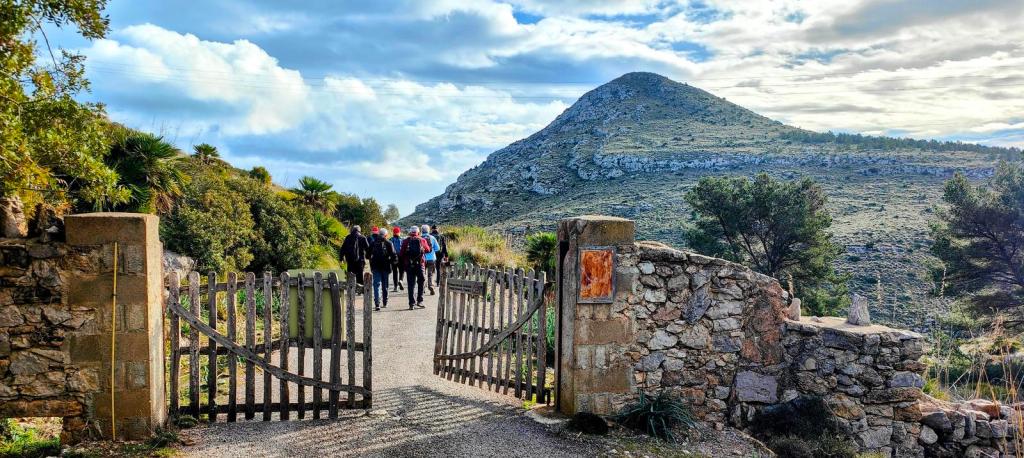 Grupo de personas caminando hacia un sendero montañoso, pasando por una puerta de madera en una cercanía de piedra.