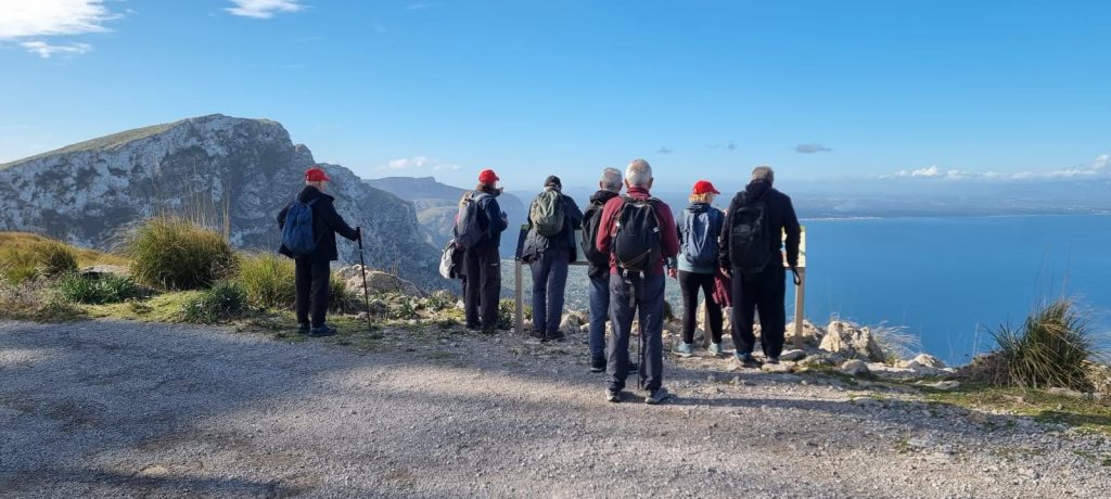Grupo de personas caminando por un sendero montañoso con vistas al mar, en un día claro y soleado, con montañas de fondo.