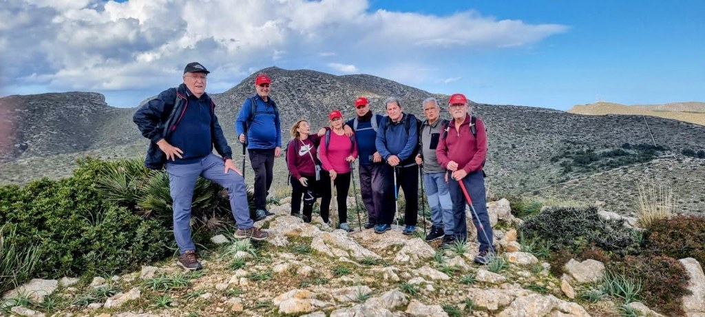Grupo de senderistas en la cima de una montaña, con vistas panorámicas de paisajes naturales. Todos llevan gorros rojos y algunos sostienen bastones de trekking. El cielo está parcialmente nublado.