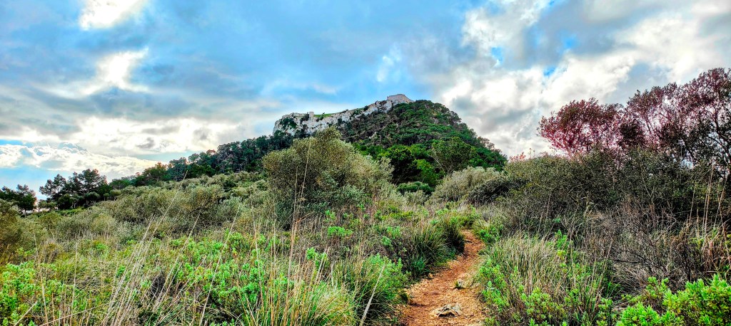 Sendero que conduce al Castillo de Santueri, rodeado de vegetación y montañas bajo un cielo nublado.