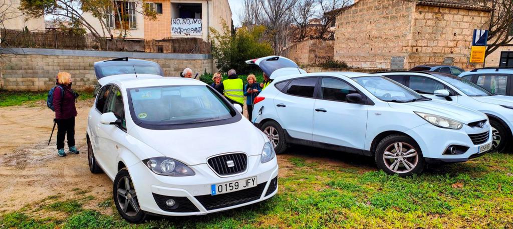 Grupo de personas en un aparcamiento, preparándose para comenzar una ruta de senderismo. Se pueden ver varios coches estacionados entre un entorno natural.