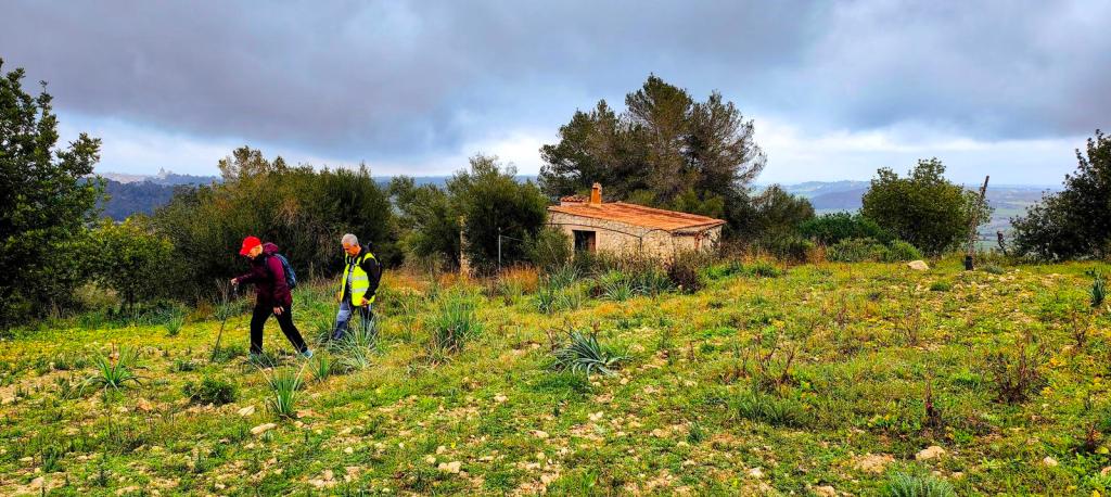 Dos personas caminando por un sendero en un paisaje rural, con una casa antigua de fondo y un cielo nublado.