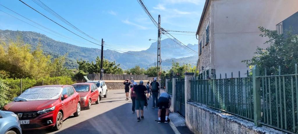 Vista de un grupo de personas caminando por una calle con coches aparcados, con montañas al fondo.