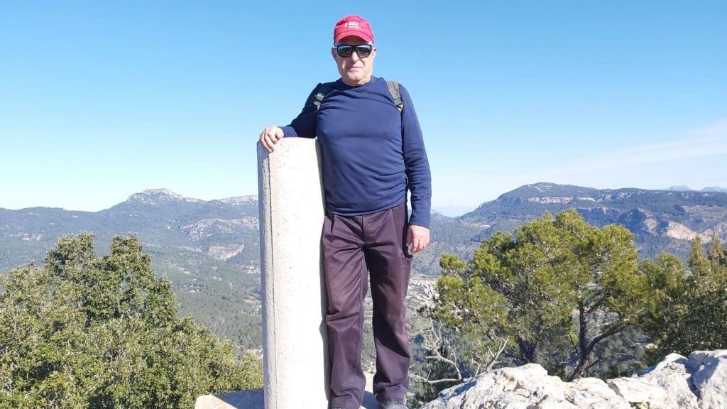 Hombre de pie junto a un vértice geodésico en la cima del Puig de Na Bauçana, con un paisaje montañoso de fondo y cielo despejado.