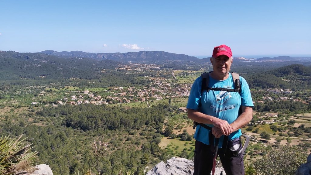 Un senderista de pie en la cima de una montaña, mirando hacia el paisaje con vistas de campos y pueblos en el fondo.