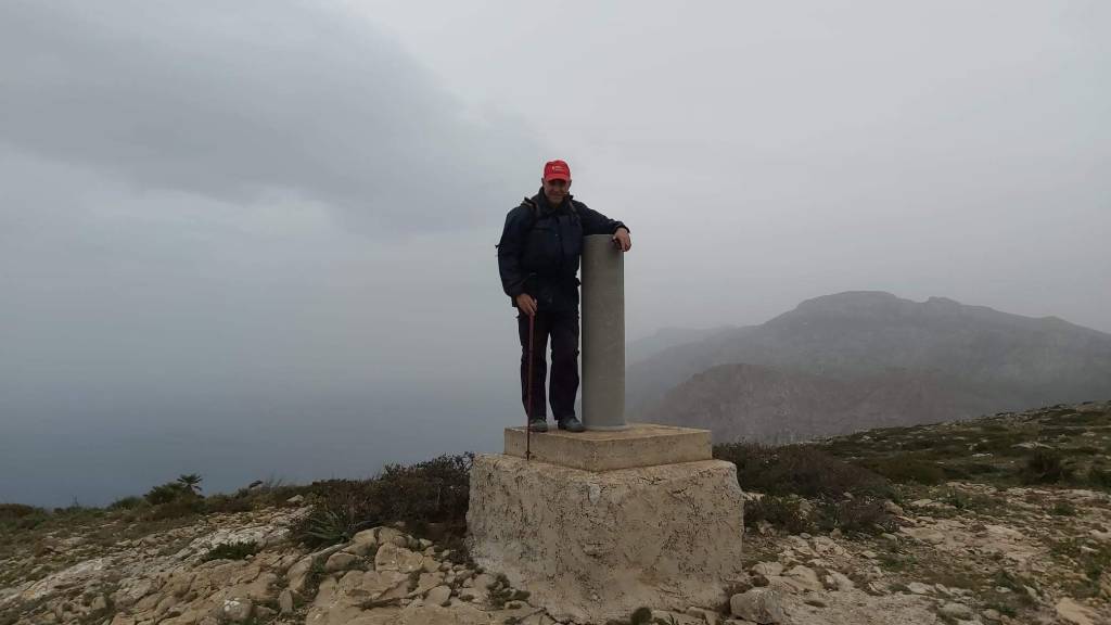 Persona de pie junto a un vértice geodésico del Puig de ses Basses en un paisaje montañoso nublado, con vegetación y vistas difusas al fondo.