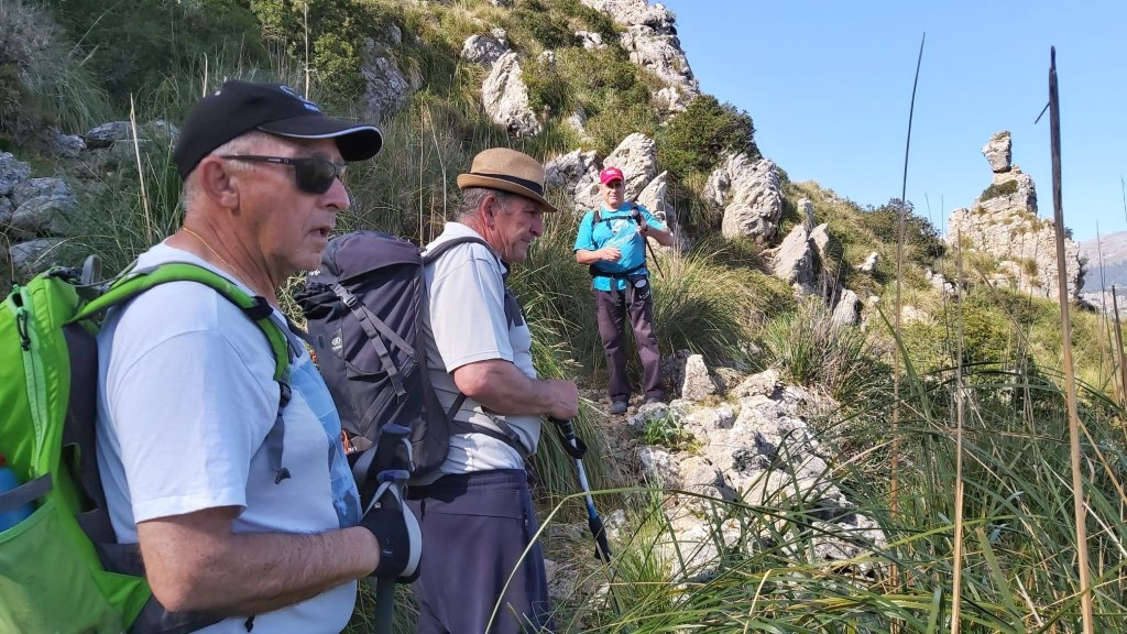 Un grupo de tres senderistas en un camino montañoso, rodeados de vegetación y rocas, con un fondo de paisaje natural.