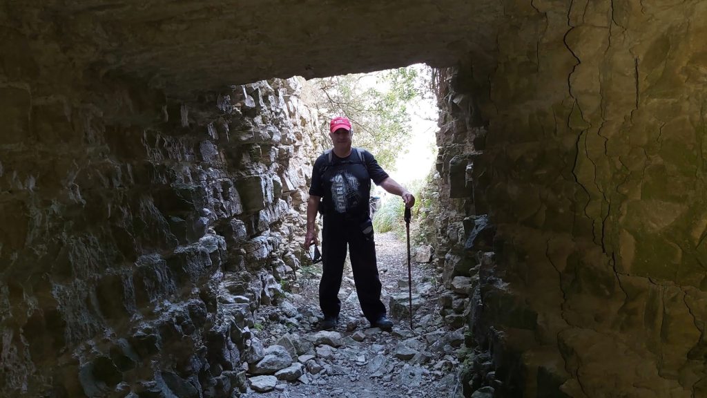 Hombre con bastón de senderismo de pie dentro de un túnel de piedra, observando hacia la salida iluminada.