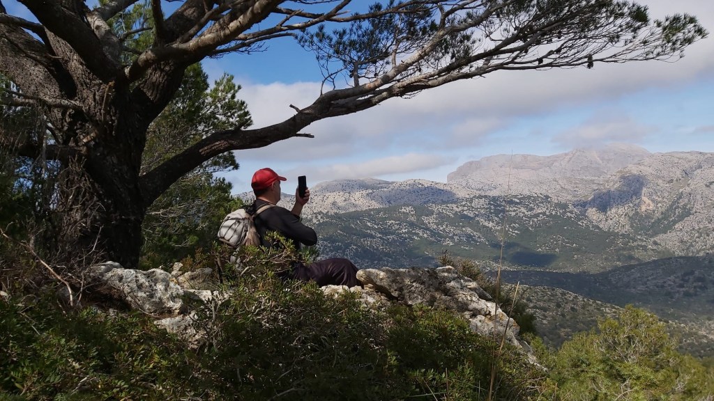 Un senderista sentado en una roca, observando el paisaje montañoso mientras sostiene su teléfono móvil. En el fondo, se pueden ver verdes colinas y montañas bajo un cielo nublado.