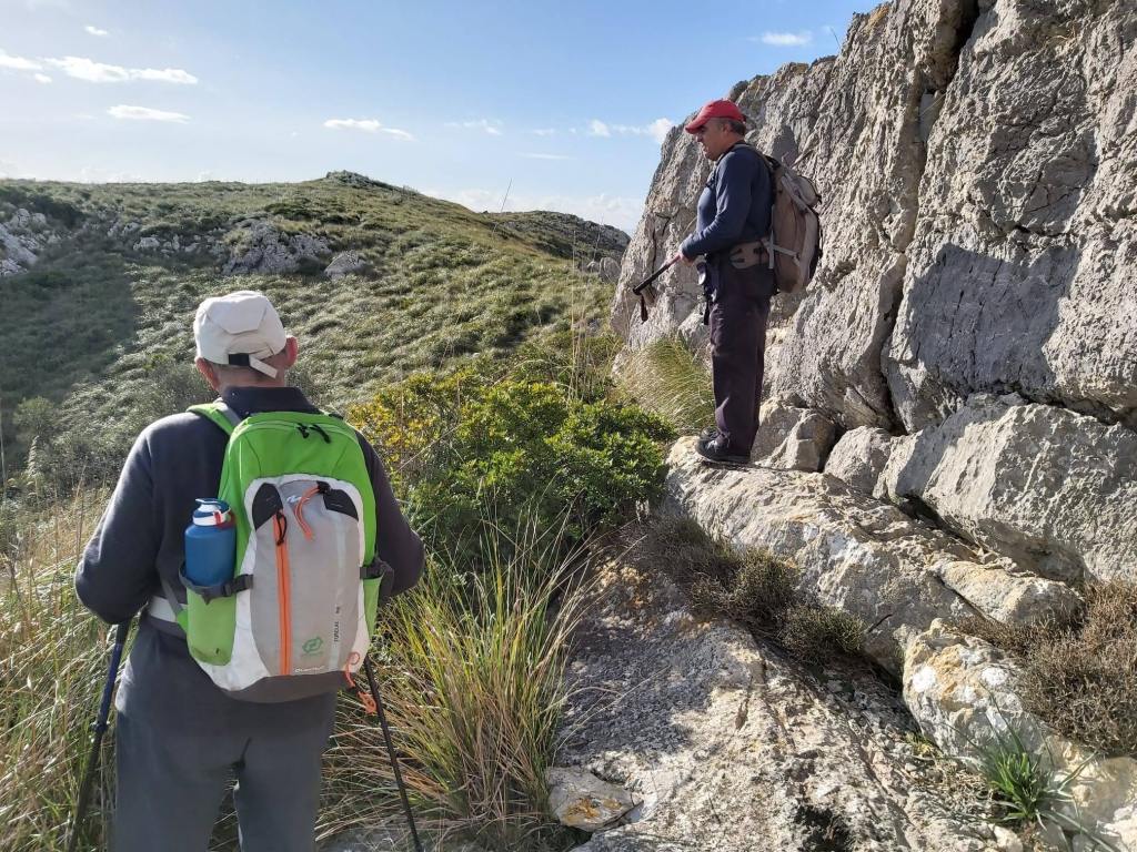 Ruta de la Sierra de Son Fe-Caminando por Mallorca Dos senderistas en un camino montañoso, rodeados de vegetación y rocas, uno de ellos sosteniendo un bastón de trekking.