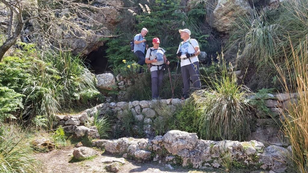 Tres excursionistas de pie en un sendero rodeado de vegetación, con un fondo rocoso y un pequeño claro en el paisaje.