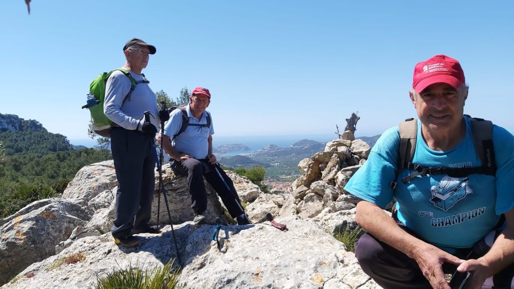 Tres senderistas descansando en una roca en la cima del Puig de Son Monjo, con vistas al paisaje montañoso y costero de fondo.