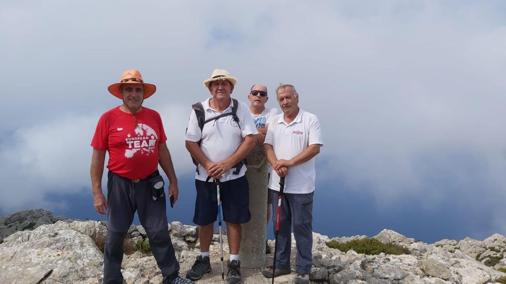 Puig Gros de Ternelles por Camí de Llinars-Caminando por Mallorca Grupo de cuatro excursionistas posando en la cima del Puig Gros de Ternelles, con vista de nubes y paisaje montañoso al fondo.