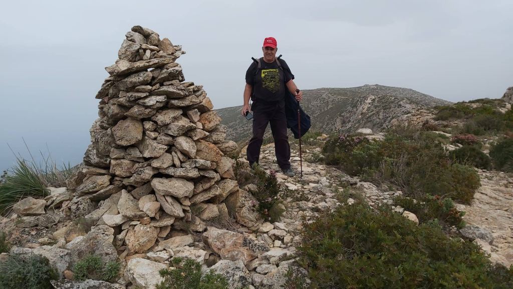 Senderista junto a un hito de piedras en un paisaje montañoso, con un fondo de colinas y vegetación mediterránea.