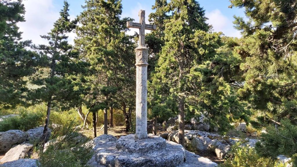 Cruz de piedra en un entorno natural, rodeada de árboles y rocas.