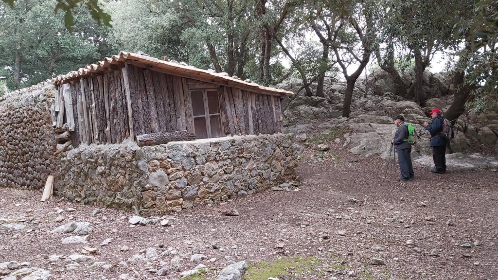 Puig del Coll del Jou desde Orient-Caminando por Mallorca Una caseta de refugio construida con paredes de piedra y madera, situada en un entorno boscoso, con dos personas de pie observando.