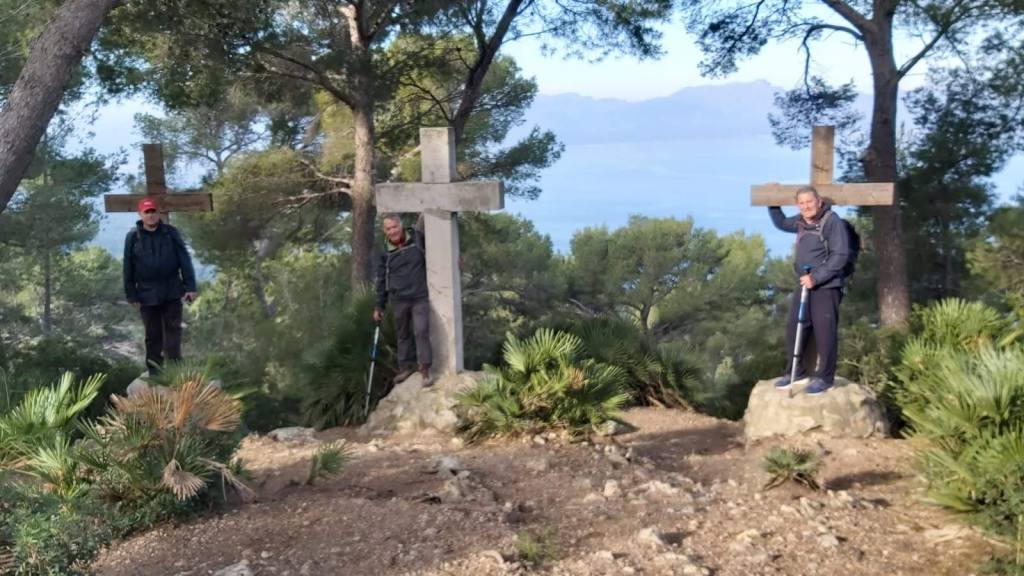 Tres hombres de pie junto a grandes cruces de madera en un área boscosa, con vistas a un paisaje montañoso y mar al fondo.