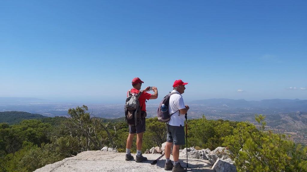 Dos senderistas en la cima del Penyal d’ Honor uno tomando fotos mientras el otro observa el paisaje, con vistas panorámicas de un valle y el cielo despejado.