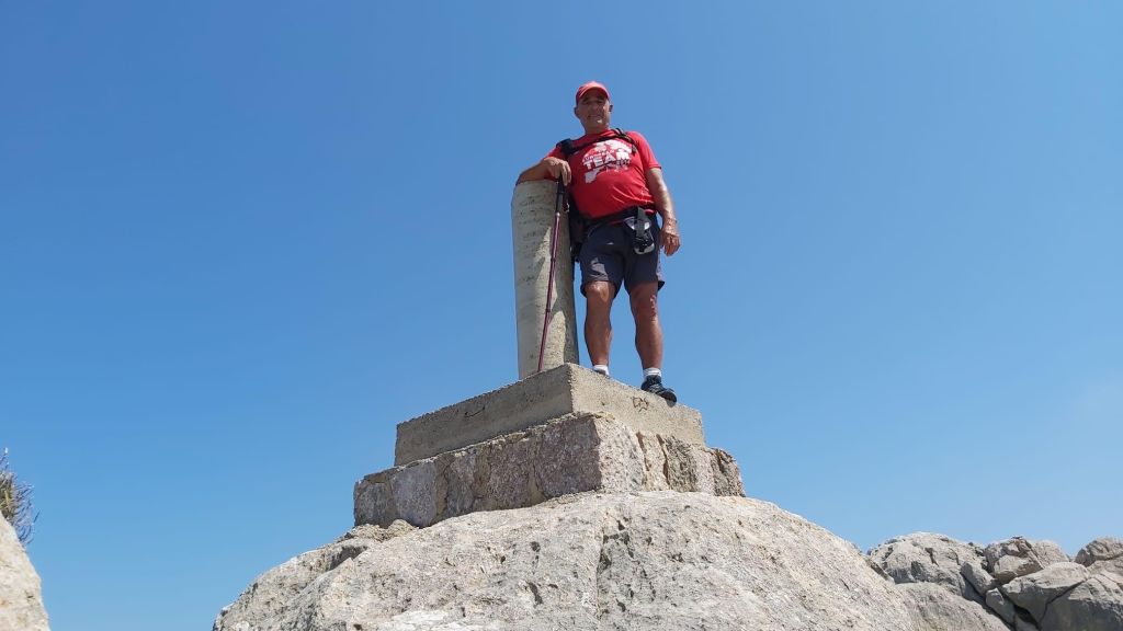 Hombre de pie sobre una roca en la cima del Puig del Morral, con un vértice geodésico detrás y un cielo azul claro.