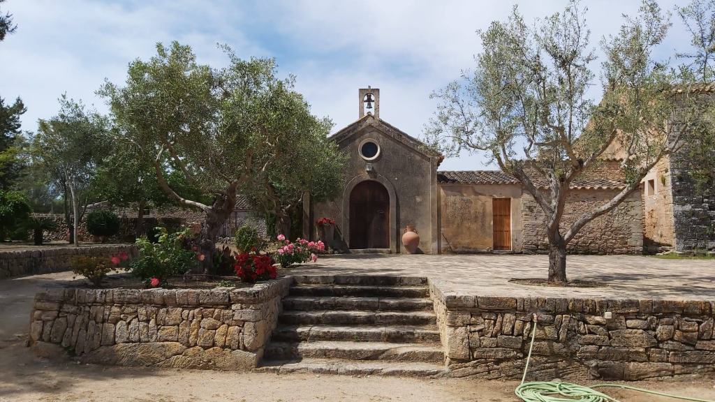 Vista de la entrada de un edificio histórico en Son Real, rodeado de árboles y flores, con escaleras de piedra que conducen a la puerta principal.