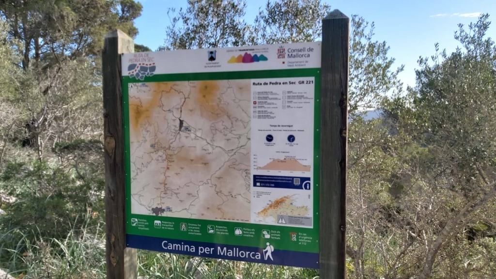 Panel informativo sobre la ruta de senderismo Puig de Na Bauçana, con un mapa y datos técnicos, rodeado de vegetación