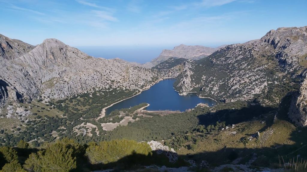 Panorámica del embalse de gorg blau rodeado de montañas en Mallorca, con un cielo despejado y vegetación en las laderas.