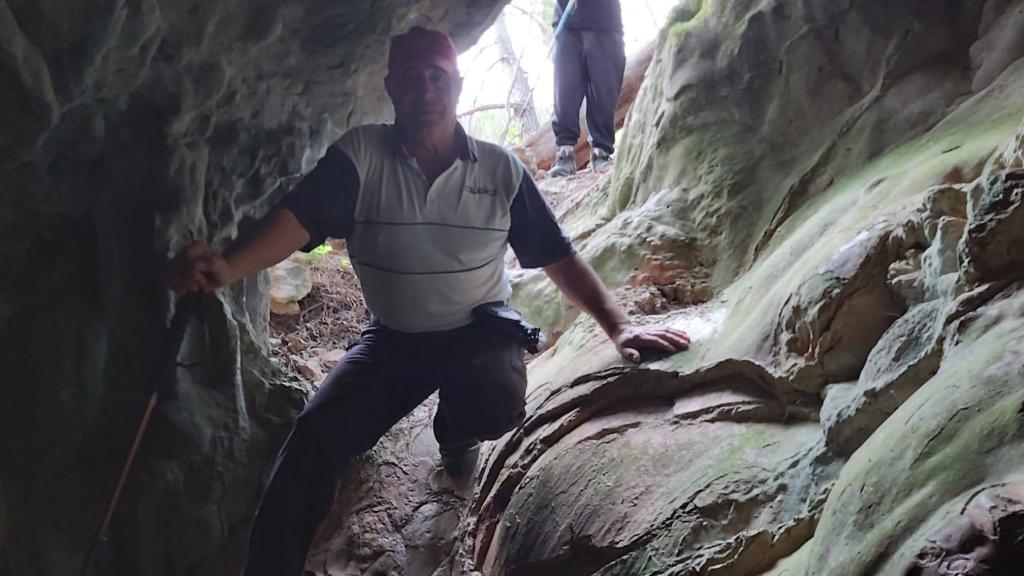 Hombre sentado en la entrada de la Cova de Can Sión, sosteniendo un bastón, con la roca verde y texturizada de fondo.