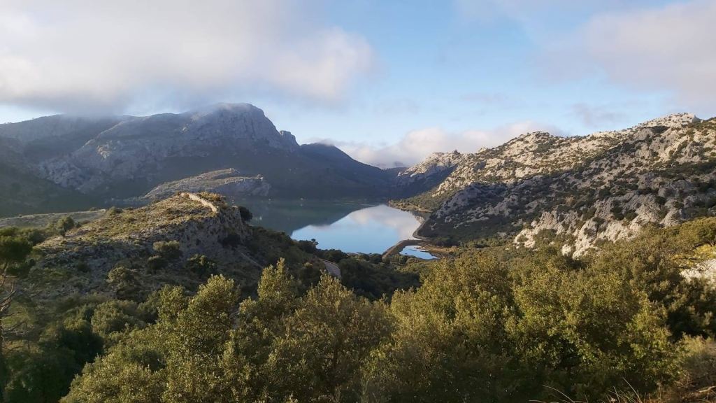 Panorámica del embalse de Cùber rodeado de montañas y vegetación, con nubes dispersas en el cielo.